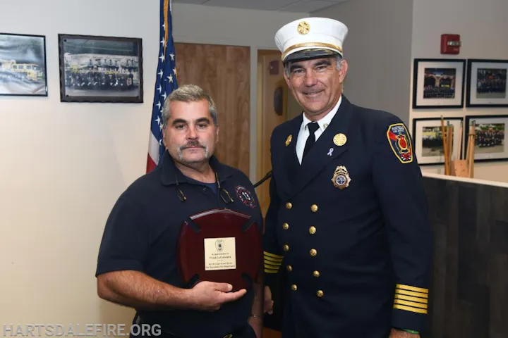 Two men in uniforms, one holding a plaque, standing together in a room with framed photos and a U.S. flag.