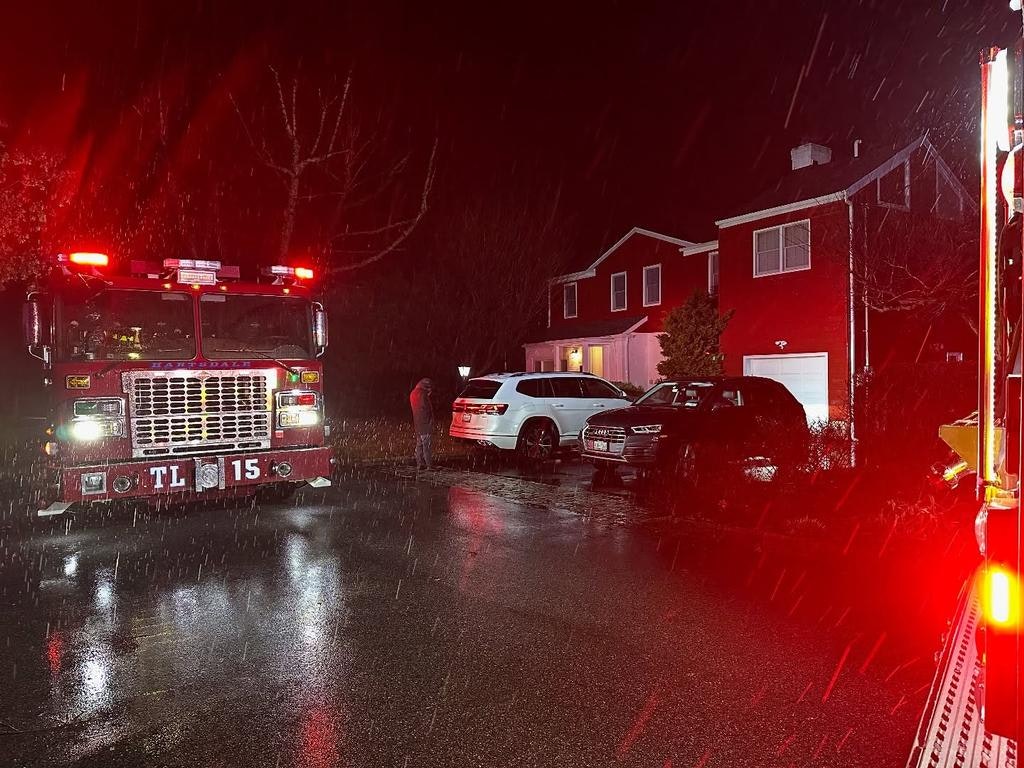 A fire truck with flashing lights is parked in a residential area at night, amid rain, near several vehicles and a red house.