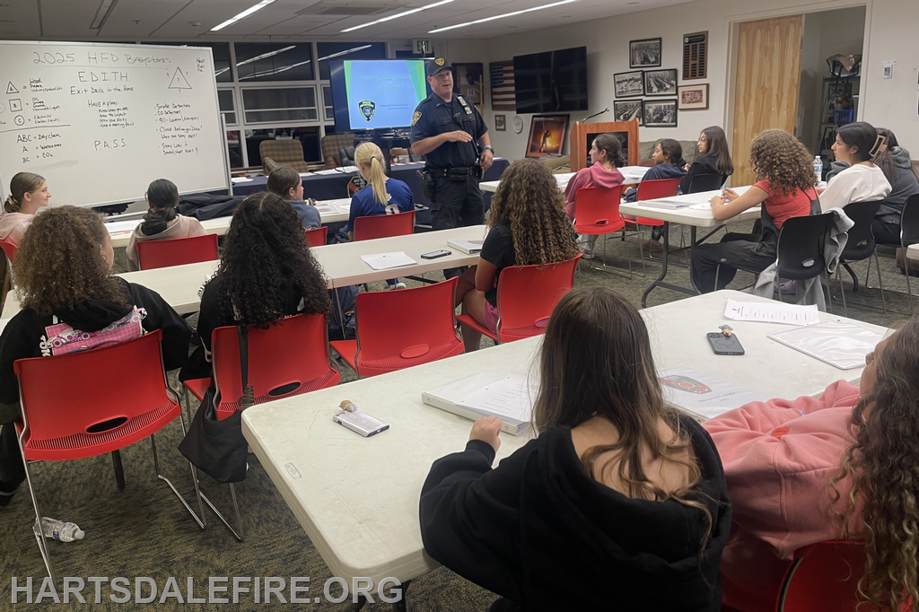 A police officer teaches a group of children about safety protocols in a classroom setting, with notes and a presentation visible.