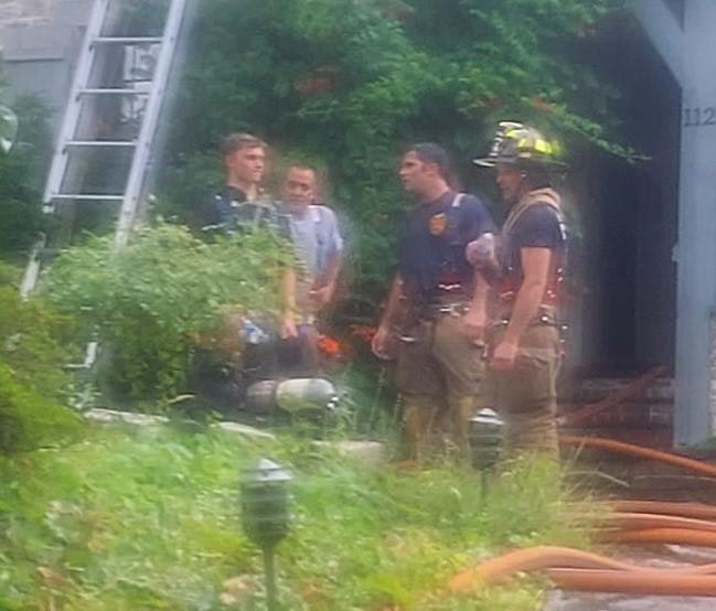 Firefighters in gear stand near a building entrance with a ladder and hoses visible, surrounded by greenery.