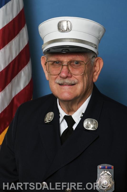 A smiling man in a fire service uniform, with a cap and badge, stands in front of an American flag backdrop.