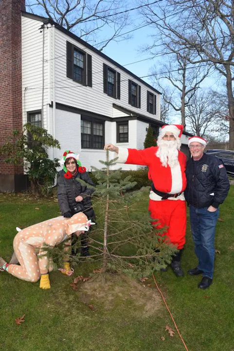 Two people in holiday attire, one as Santa, pose with a small tree and someone in a reindeer costume in front of a house.