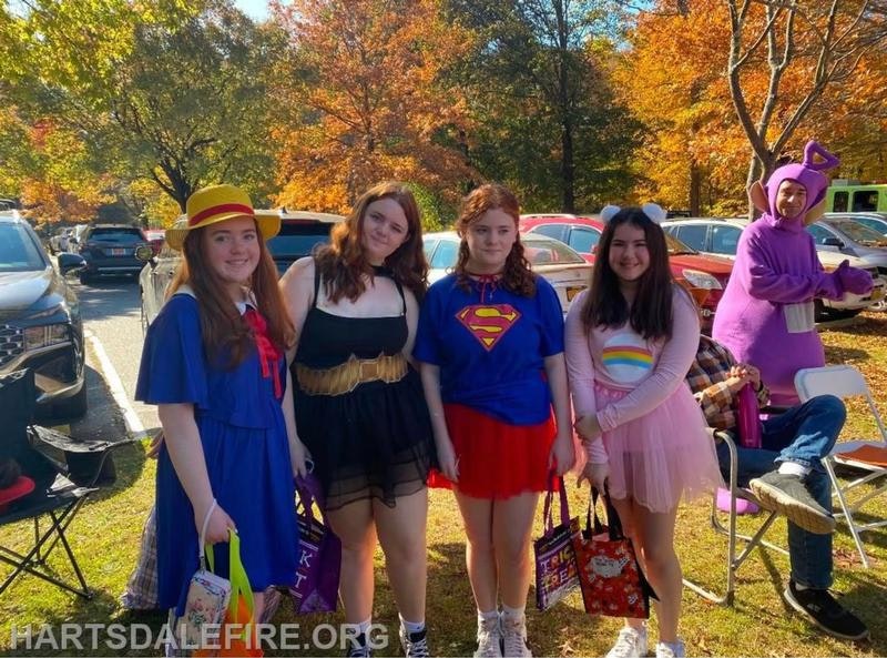 A group of kids dressed in colorful costumes for a celebration, surrounded by autumn foliage and cars.