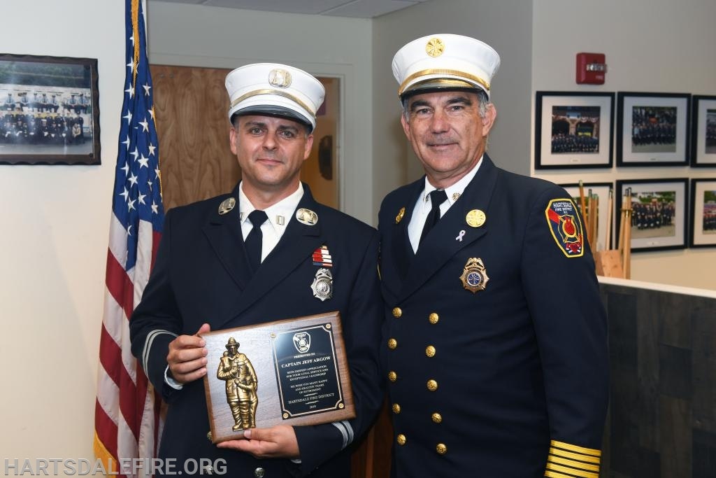 Two uniformed firefighters, one holding a plaque, stand next to an American flag in an office setting.