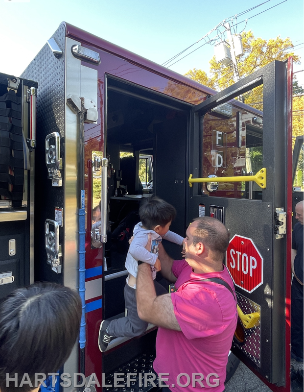 A firefighter helps a child climb into a fire truck while others watch nearby.