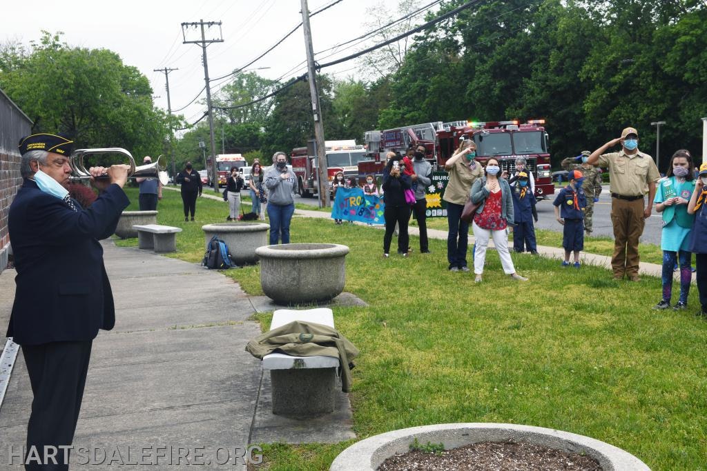 A man plays a bugle while people, including scouts, salute outdoors near fire trucks.