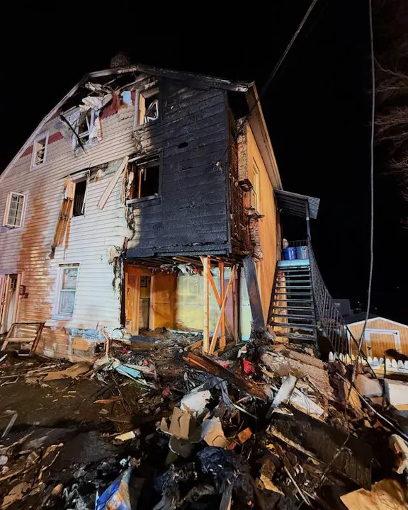 The image shows a house severely damaged by fire, with charred walls and debris scattered around the base.