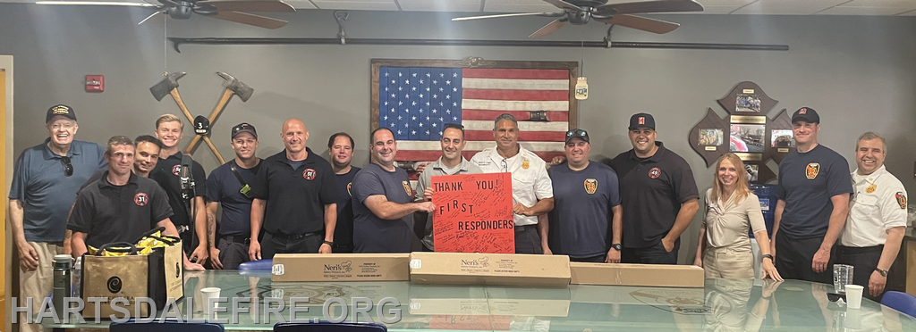 A group of people, including firefighters and officials, poses with a "Thank You, First Responders" sign in front of an American flag.