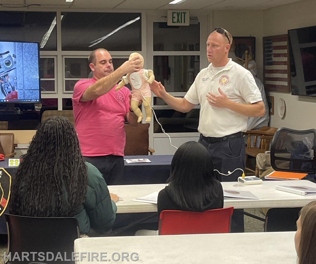 A man in a pink shirt holds a doll while another man in a fire department uniform explains something to a group in a classroom.