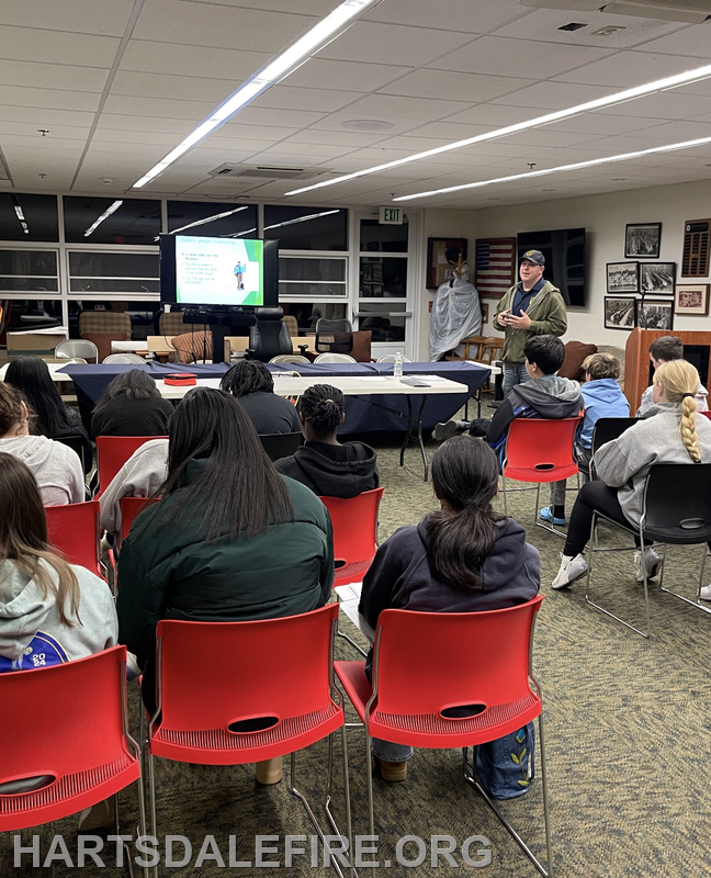 A speaker presents to a group in a classroom setting, with a screen displaying information. Audience members are seated.