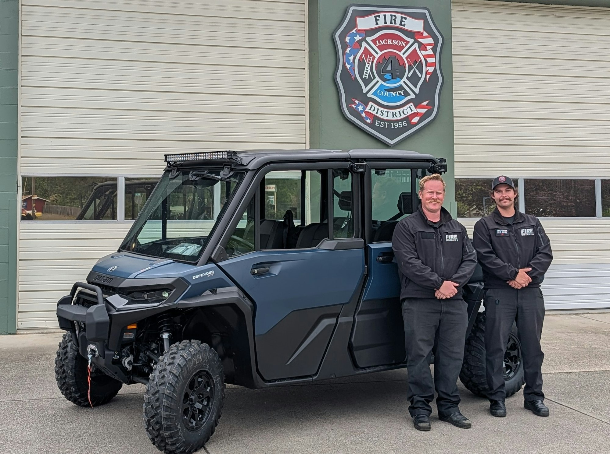 Two firefighters stand beside a blue utility vehicle in front of a fire district station, showcasing their equipment and teamwork.