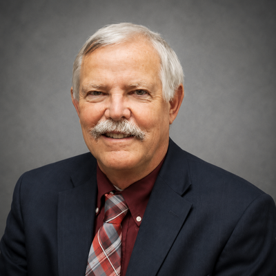 A man with a mustache, wearing a suit and tie, smiles in front of a gray background.