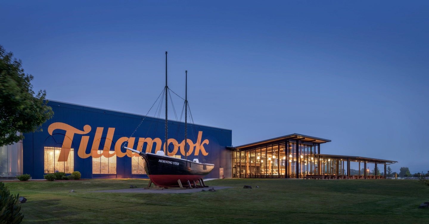 A boat outside a modern building with "Tillamook" in large letters, illuminated at dusk.