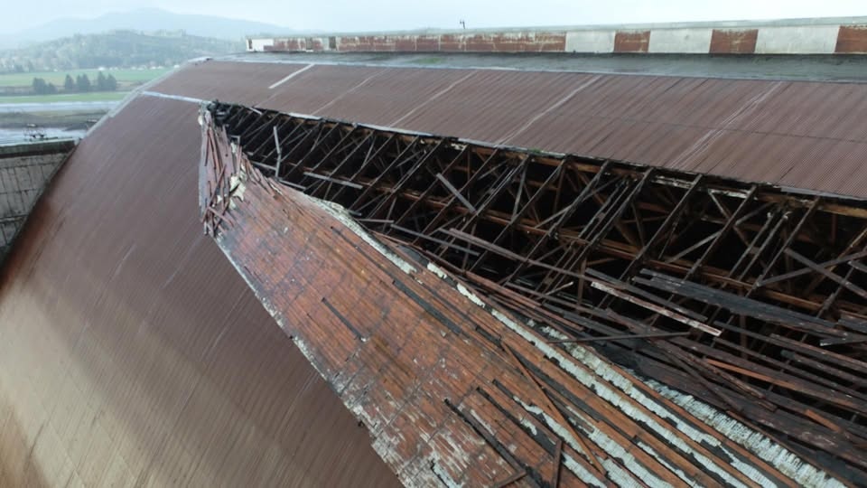 The image shows a damaged, peeling roof with exposed wooden supports and a mountainous landscape in the background.
