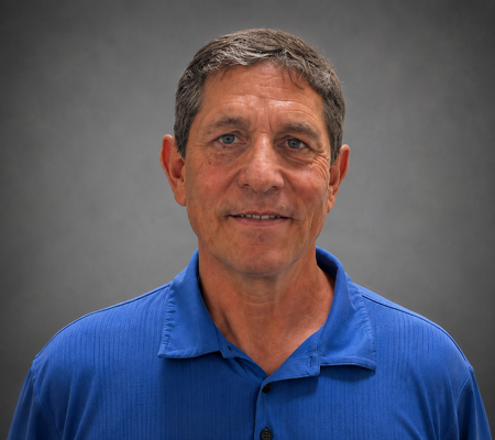 A man with short hair wearing a blue shirt, smiling against a gray background.