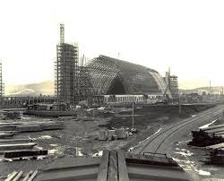 Construction site with a large arched structure being built, surrounded by scaffolding and industrial materials.
