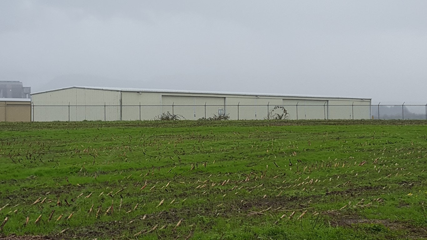 A large industrial building with a metal roof, set behind a chain-link fence, with a grassy field in the foreground under a cloudy sky.