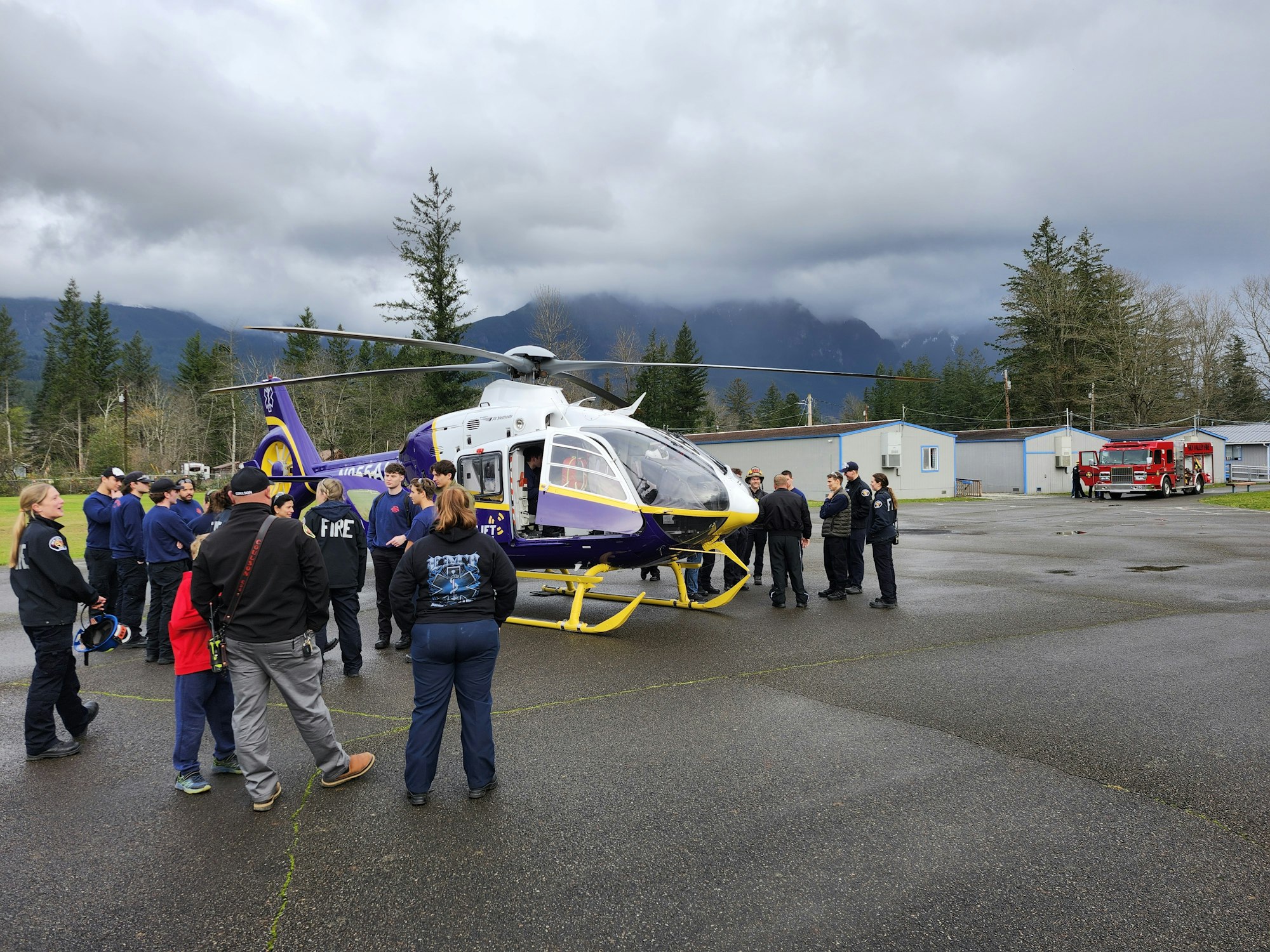 A group of people, likely first responders, are gathered around a helicopter, with a fire truck nearby and a cloudy backdrop.