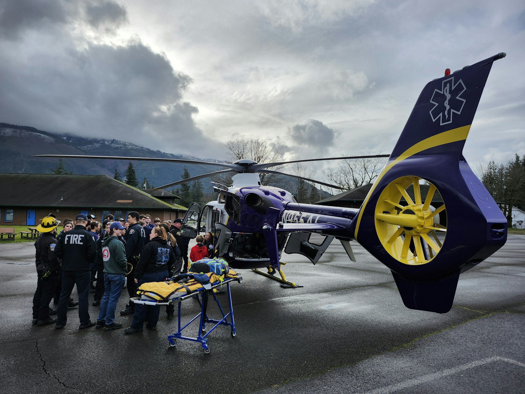 A group gathers around a purple and yellow medical helicopter, likely for a demonstration or training, with a stretcher nearby.
