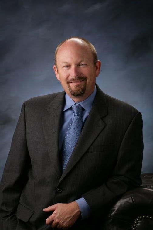 A man in a suit poses with one hand in his pocket and one resting on a dark, leather chair, against a dark background.