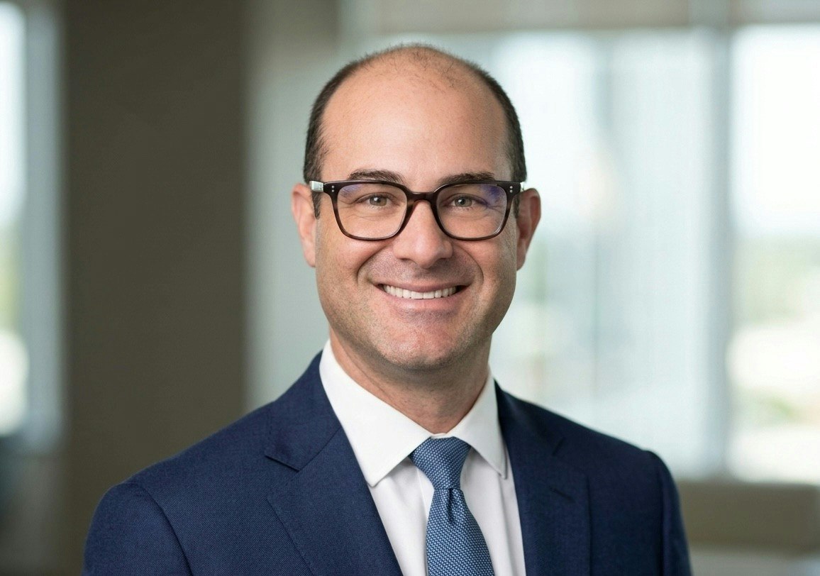 The image features a smiling man in a suit and tie, wearing glasses, with a professional background.