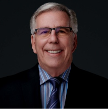A smiling man with glasses, wearing a suit and tie, against a dark background.