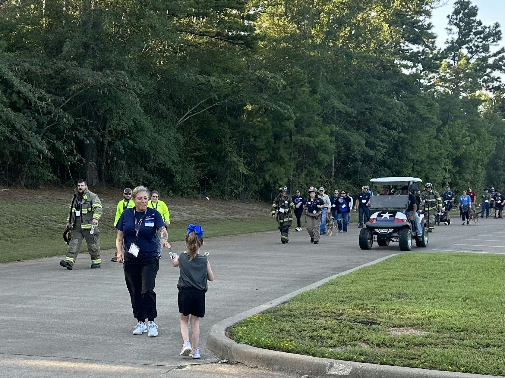 A group of people, including firefighters and a child in a blue bow, walk on a path next to a golf cart with a star on it.