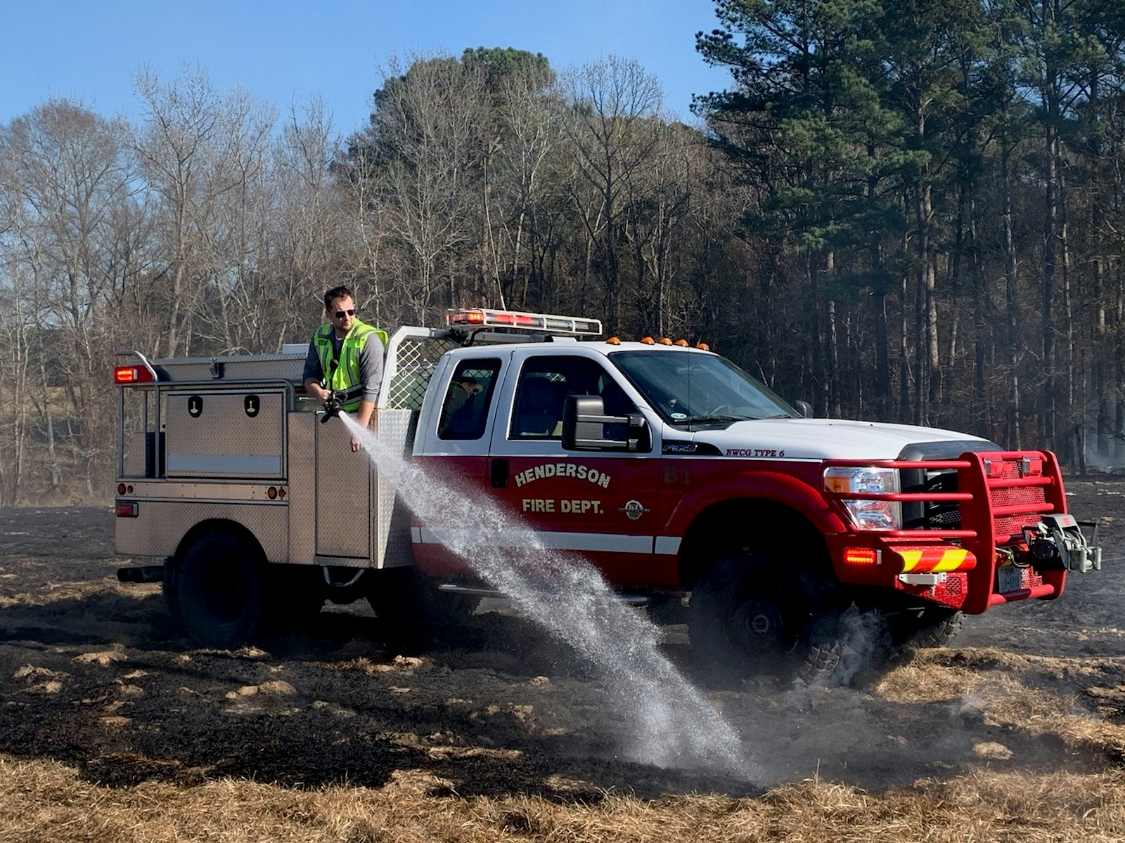Firefighter using a hose from a fire truck to spray water on a grassy area.