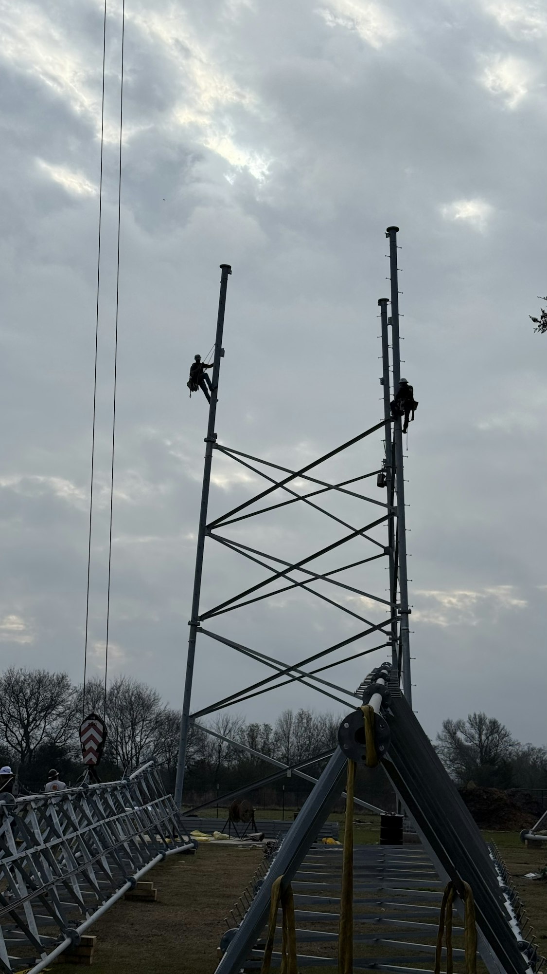 Workers climbing a tall metal structure on a cloudy day, with equipment and materials in the foreground.