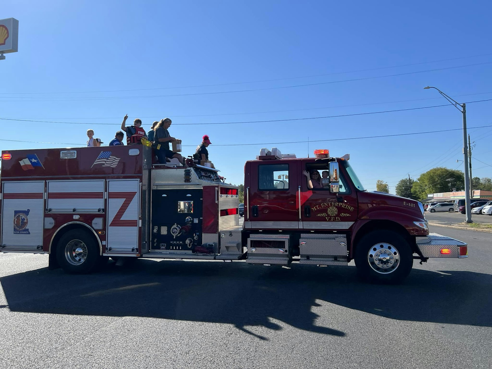 A fire truck with people aboard, likely in a parade or community event, waving to onlookers under a clear sky.