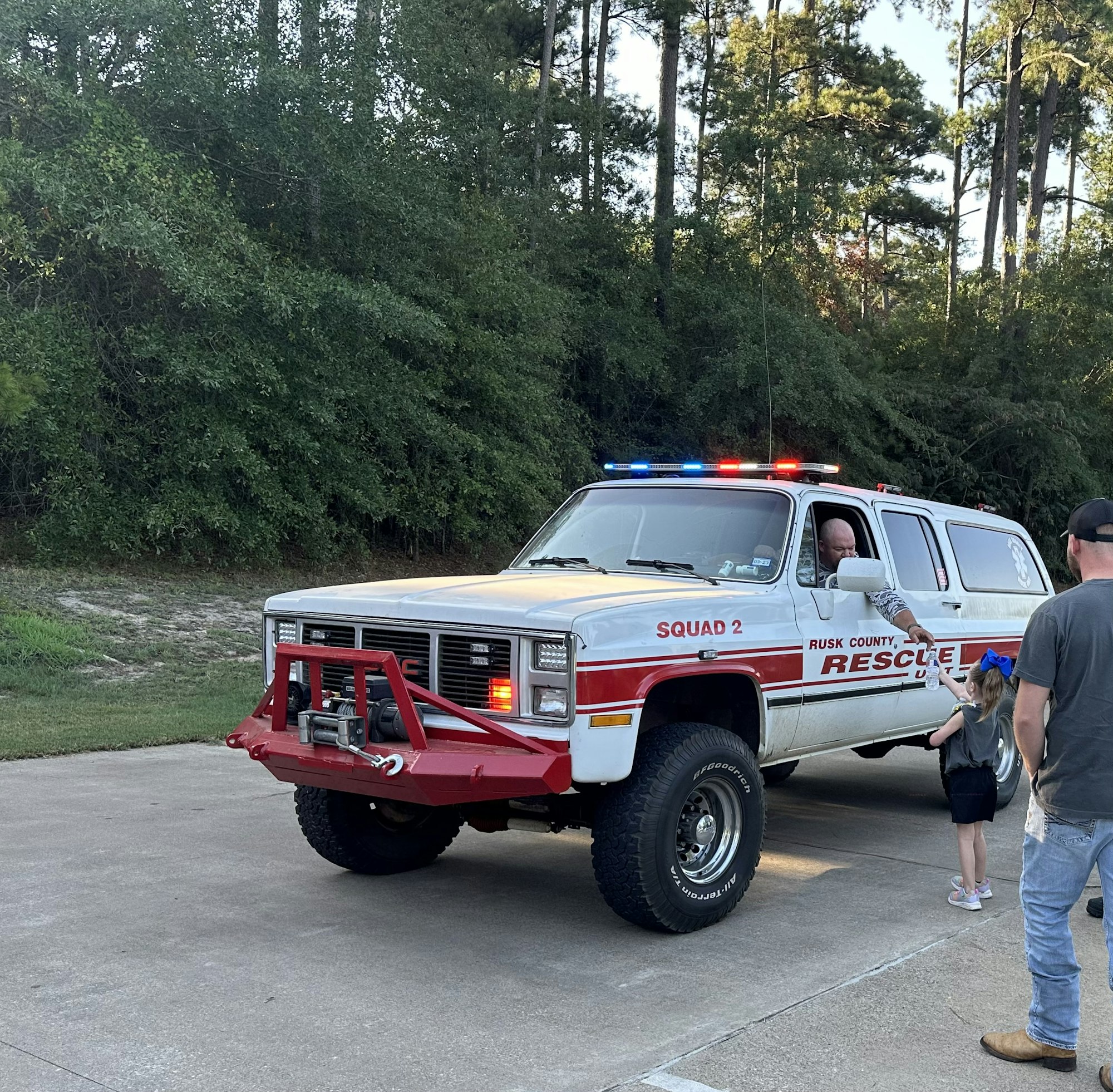 A Rusk County rescue vehicle with lights on is parked on a road, alongside several people.