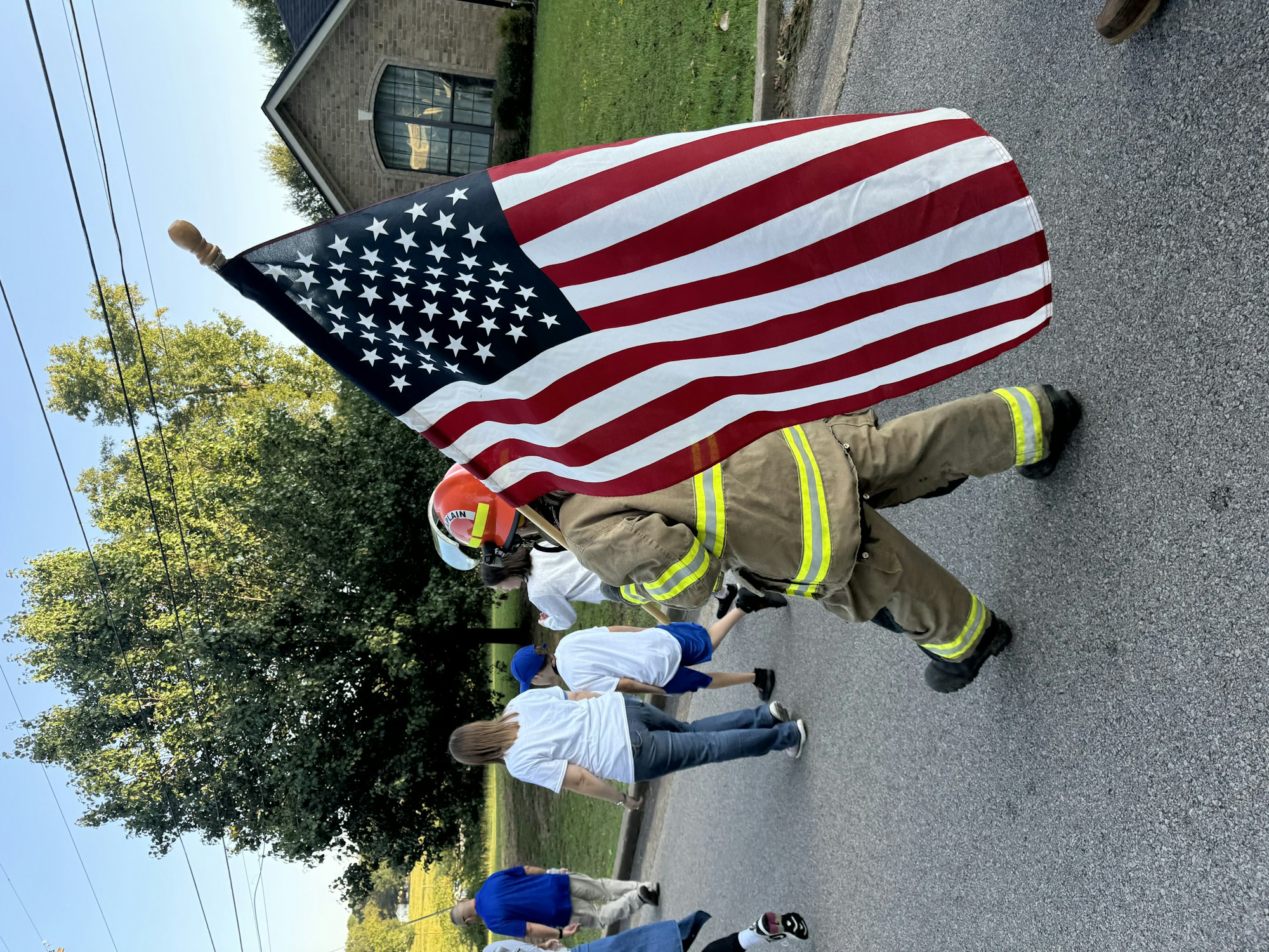 A firefighter carries an American flag during a parade, with people walking on a road.