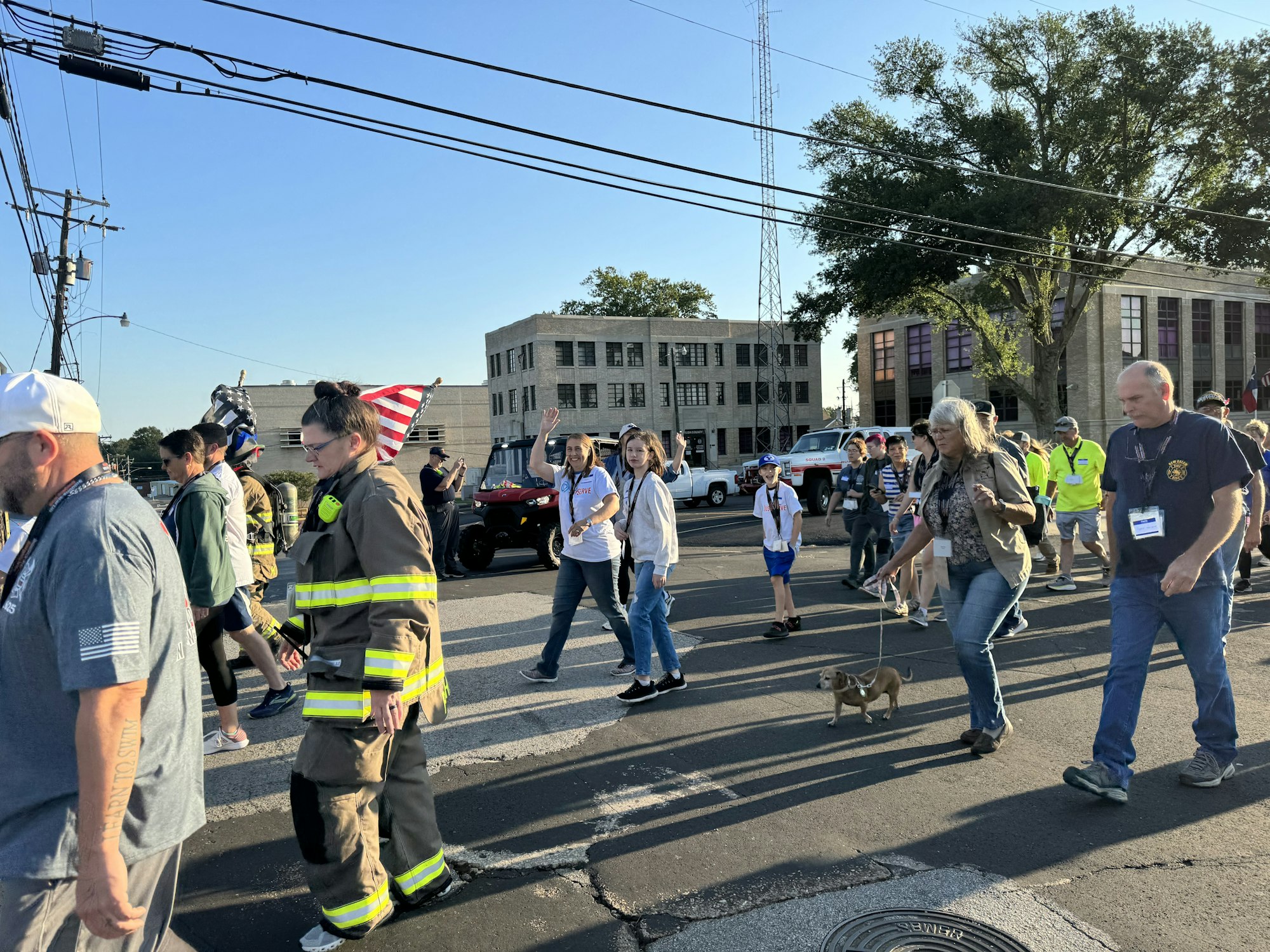 A group of people, including a firefighter in uniform and a small dog, walk down a street. Some carry American flags.