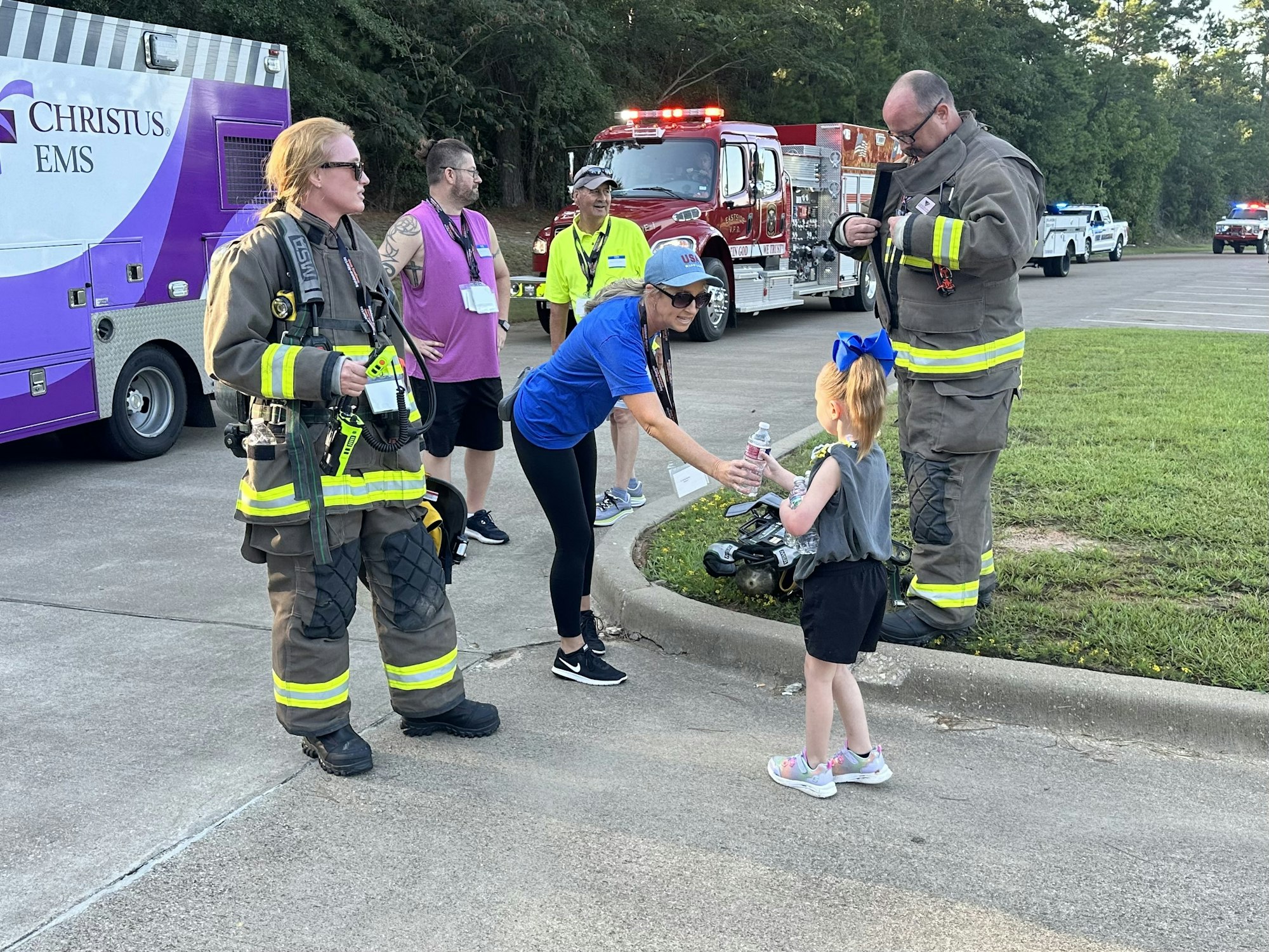 Firefighters and civilians near emergency vehicles; a woman in blue hands a water bottle to a child.