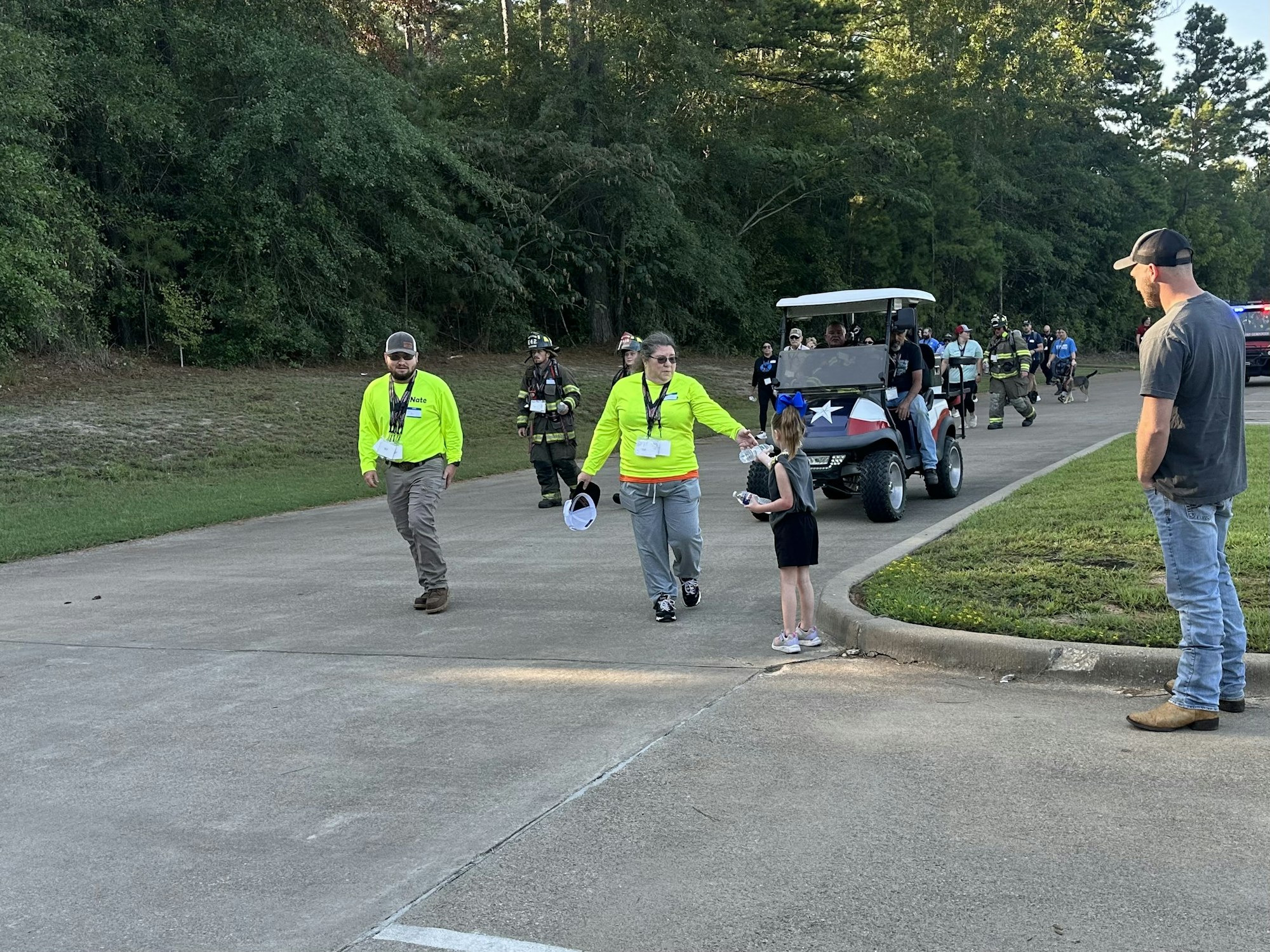 A group of people in bright shirts and uniforms walk on a path, others ride a golf cart with a Texas flag design.