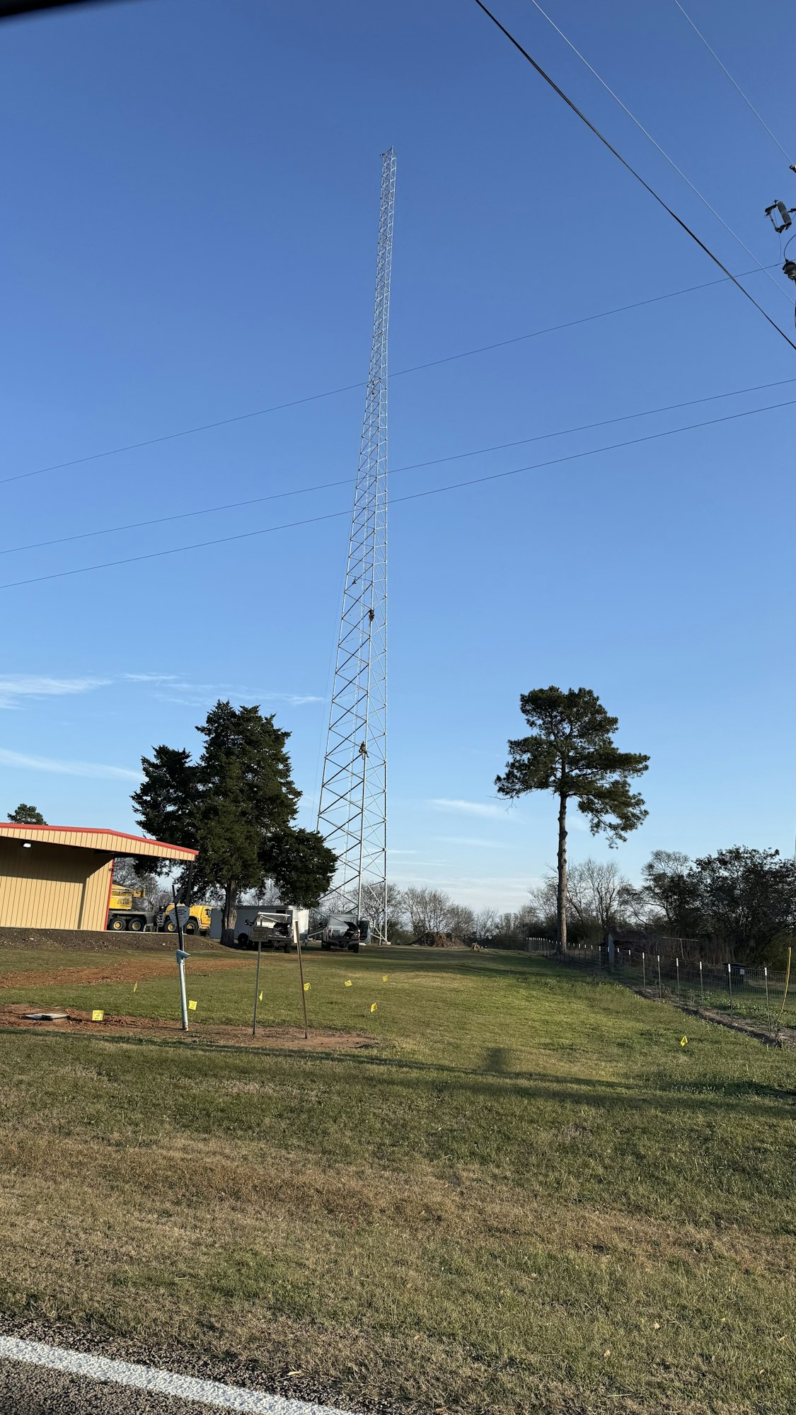 A tall radio tower stands near a building, with a clear blue sky overhead and a few trees and vehicles nearby.