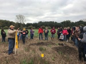 Group of people listening to a person gesturing in a grassy field, possibly an outdoor class or tour.