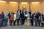 A group of people standing in a council chamber with flags and a seal, holding a framed certificate.