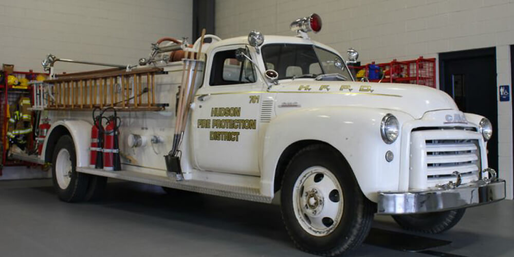 A vintage white fire truck with ladders and equipment, parked indoors, labeled "Hudson Fire Protection District."