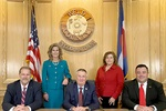 Five people, formal attire, seated and standing by flags and wooden seal on the wall.