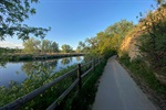 A paved path runs alongside a river, bordered by a wooden fence and trees under a clear blue sky.