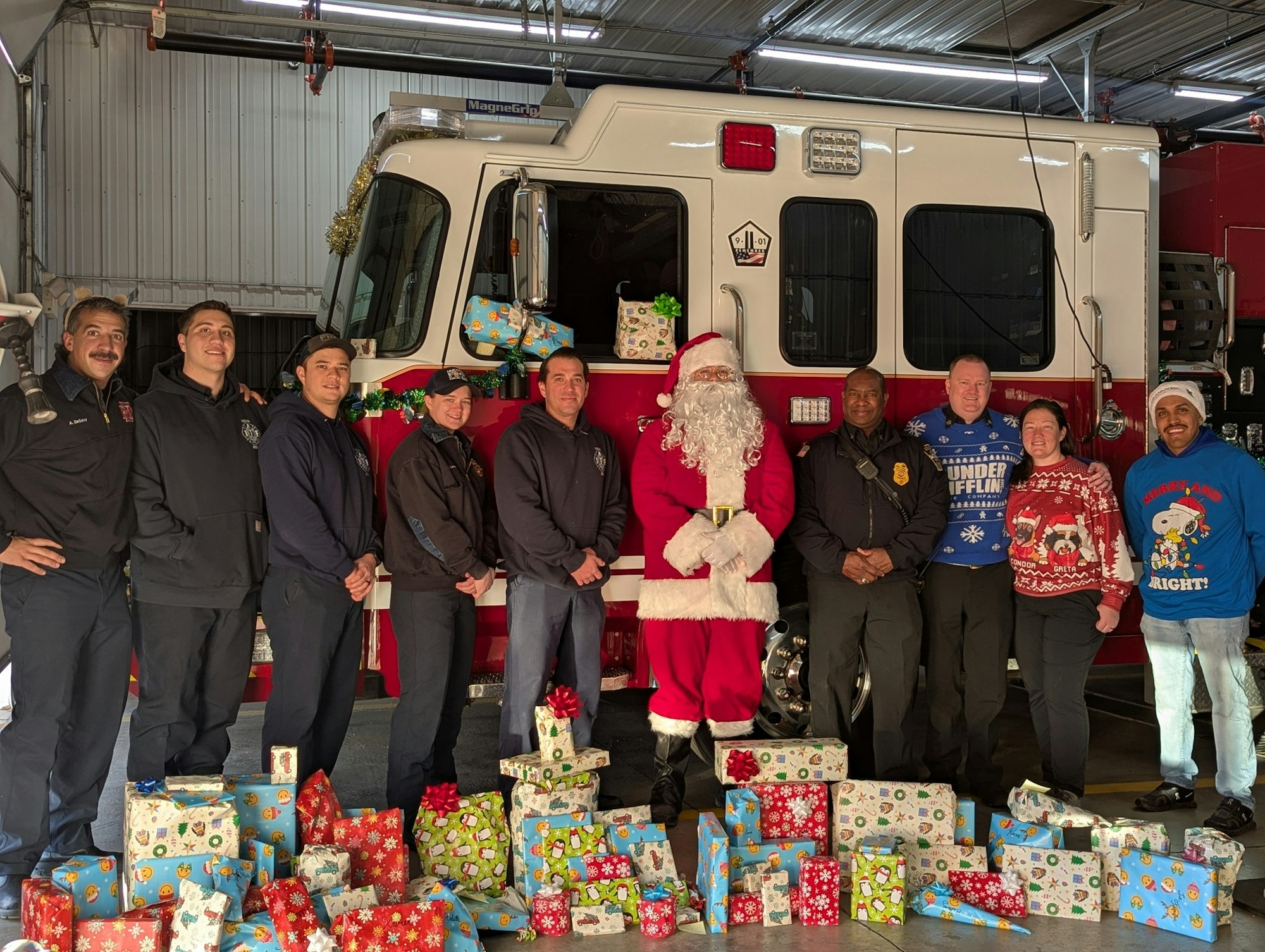 A group of firefighters and volunteers, including someone dressed as Santa, pose with festive gifts in a fire station.