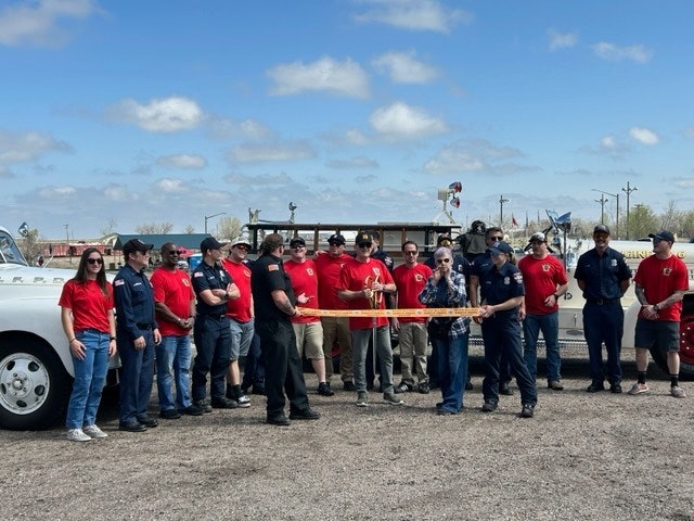 A group of people in red shirts and uniforms is gathered for a ribbon-cutting ceremony outdoors, celebrating a new opening.