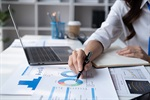 A person working on a laptop, analyzing charts and graphs with a pen, and a coffee cup nearby.