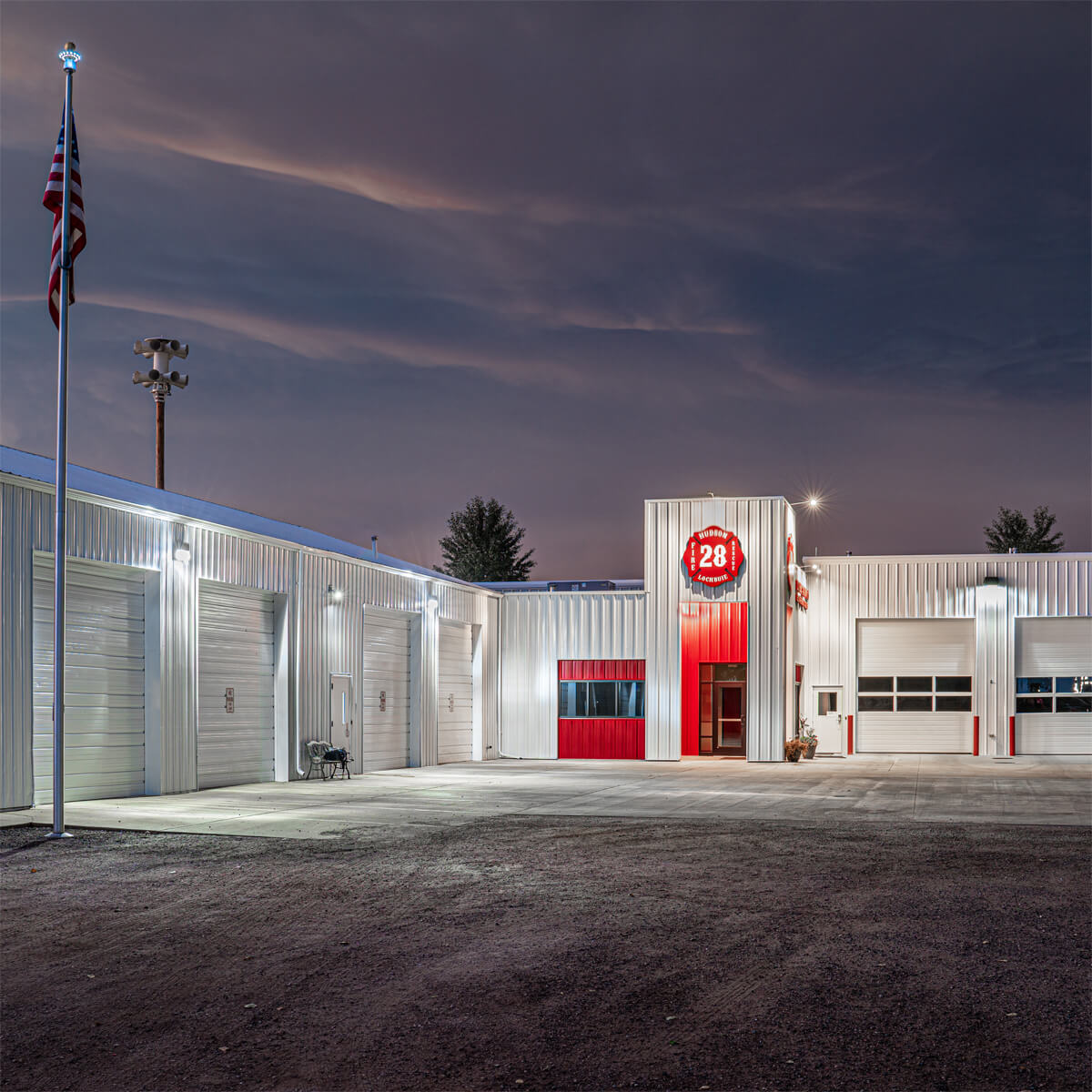 A fire station with a large number "28" sign above the entrance, white garage doors, red accents, and an American flag on a pole at dusk.
