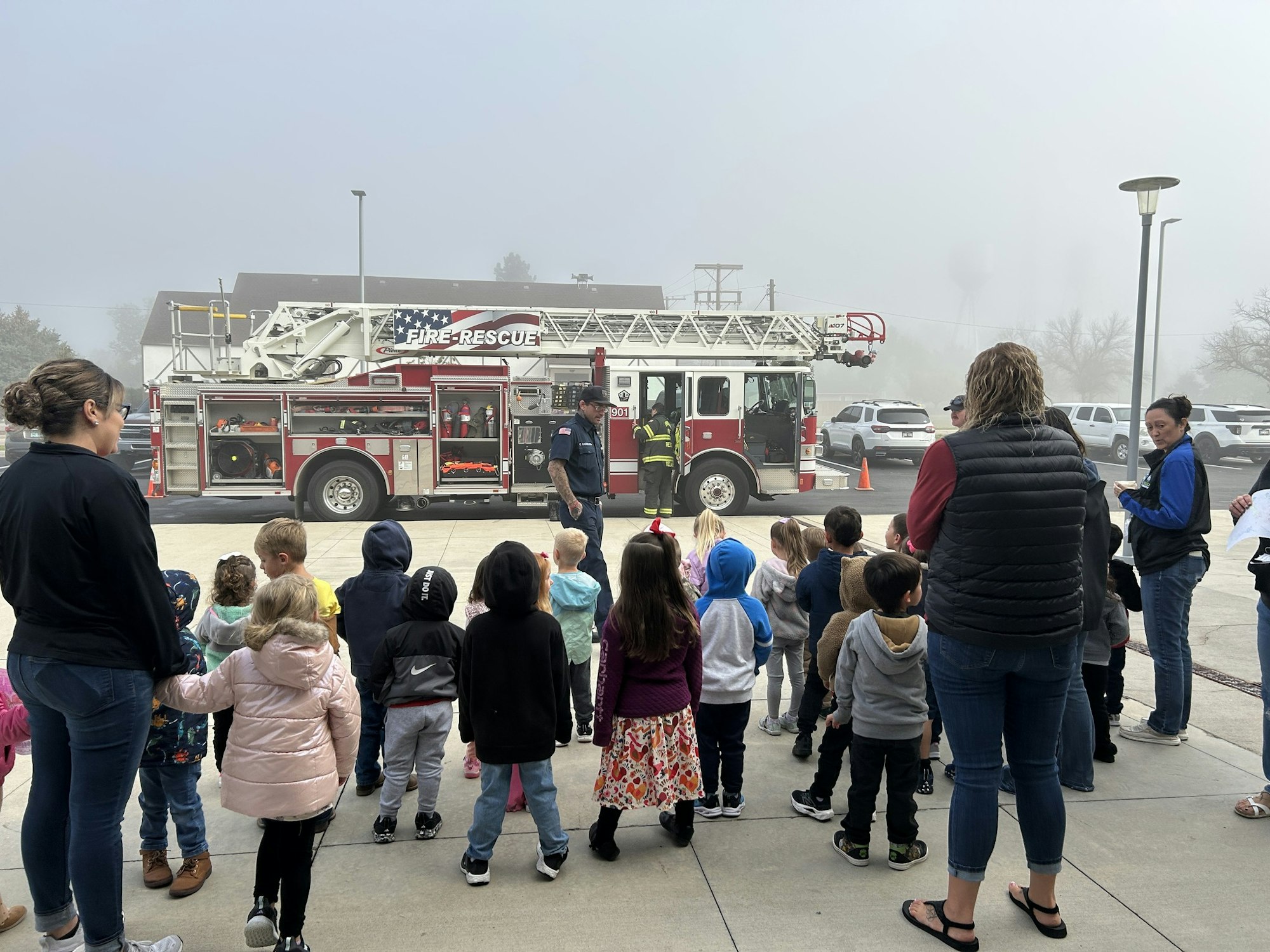 A group of children watches a fire truck demonstration on a foggy day, with adults supervising the event.