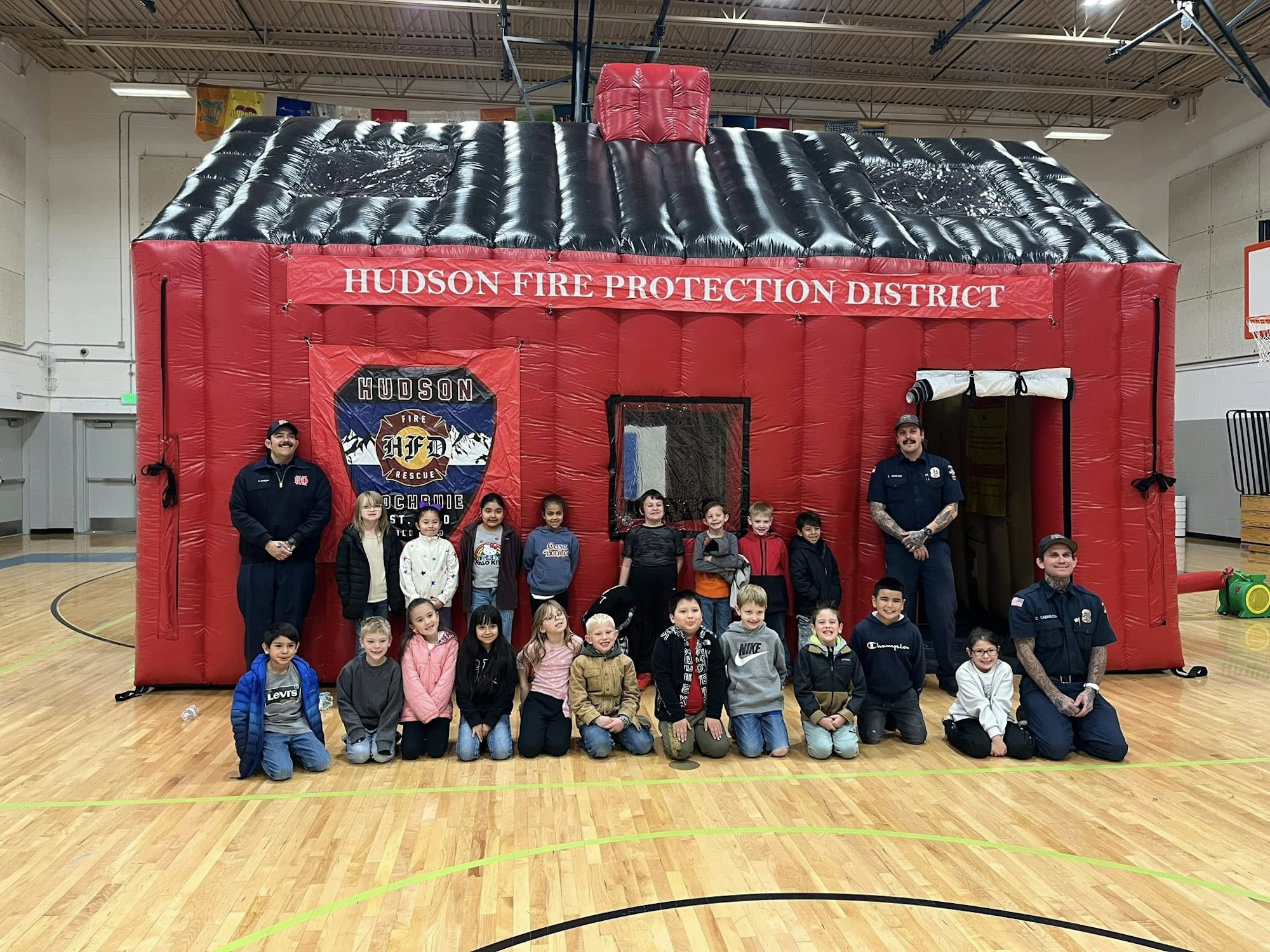 A group of children poses with firefighters in front of an inflatable firehouse in a gymnasium.