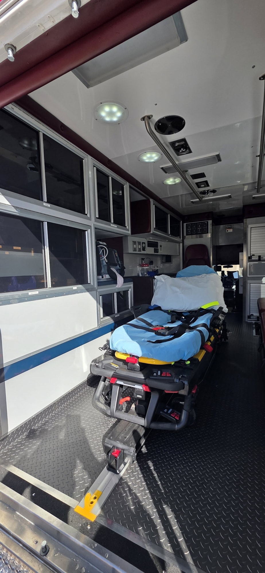 The image shows the interior of an ambulance, featuring a stretcher with blue padding, medical equipment, and storage compartments.