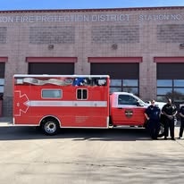 A red emergency vehicle is parked in front of a fire station, with two personnel posing beside it.