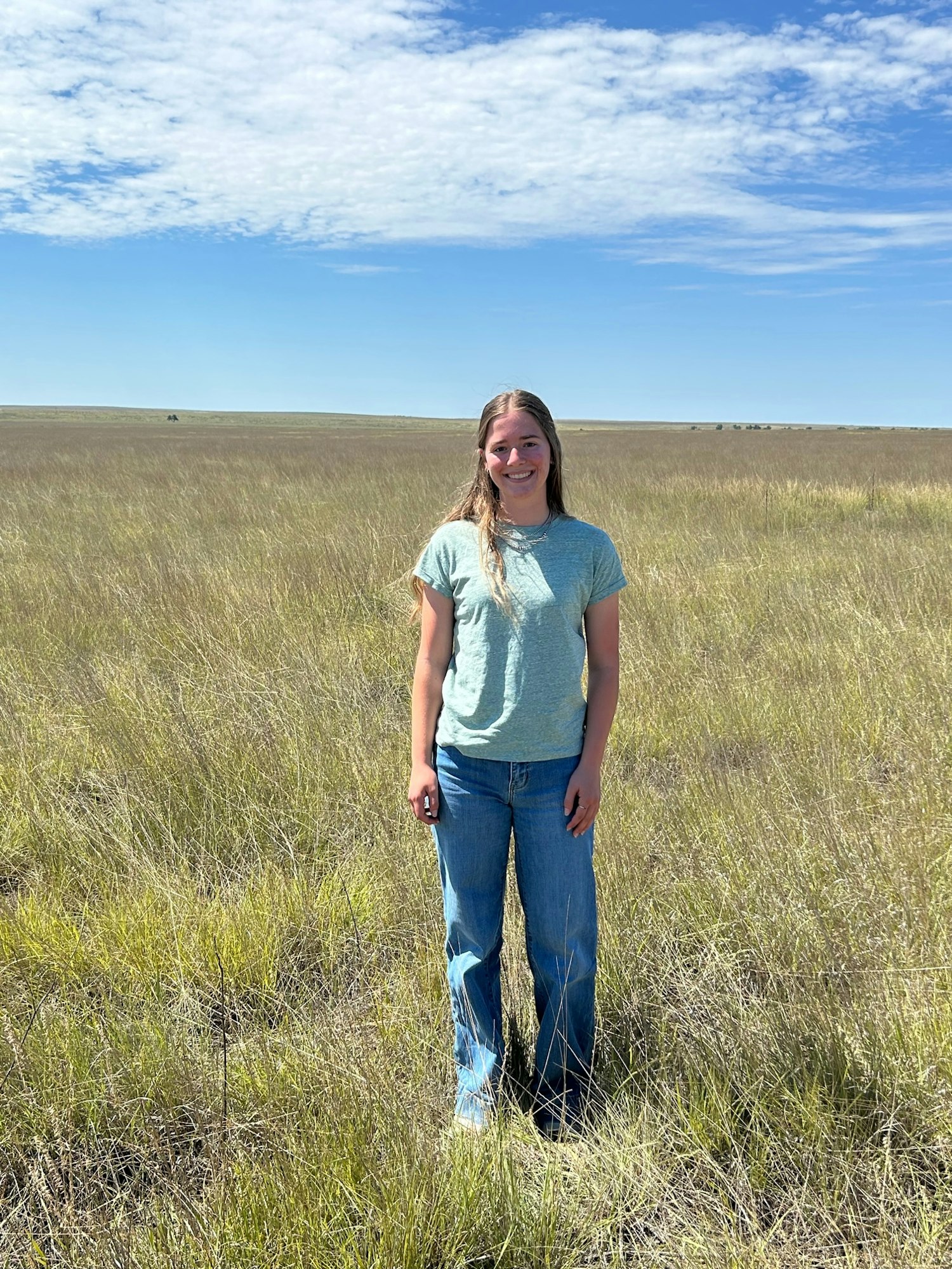 A person stands in a grassy field under a blue sky with scattered clouds, wearing a light shirt and jeans.